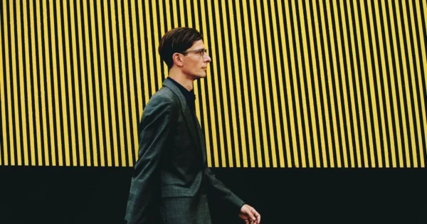 Official portrait of Mark Adam Harold walking in front of a wall with painted black and yellow stripes, photo by Arcana Femina